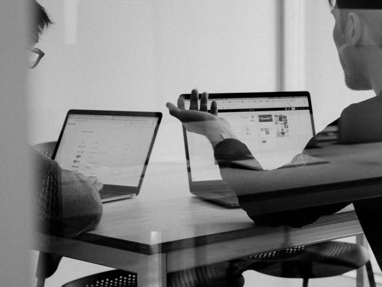 Zwei Personen arbeiten an Laptops an einem Tisch und diskutieren.